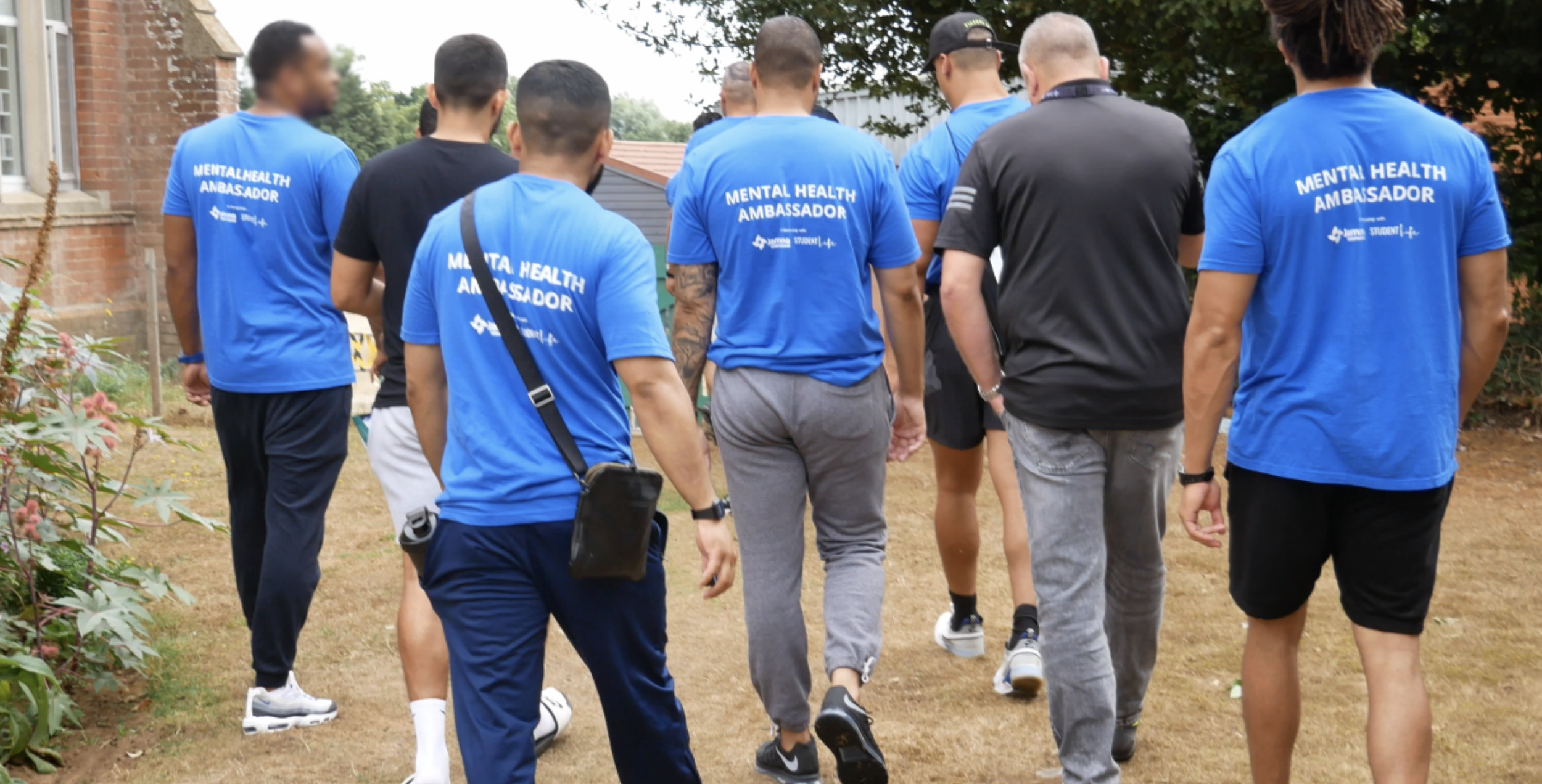 A group of men walk through a prison garden wearing blue t-shirts that read “Mental Health Ambassador” on the back.