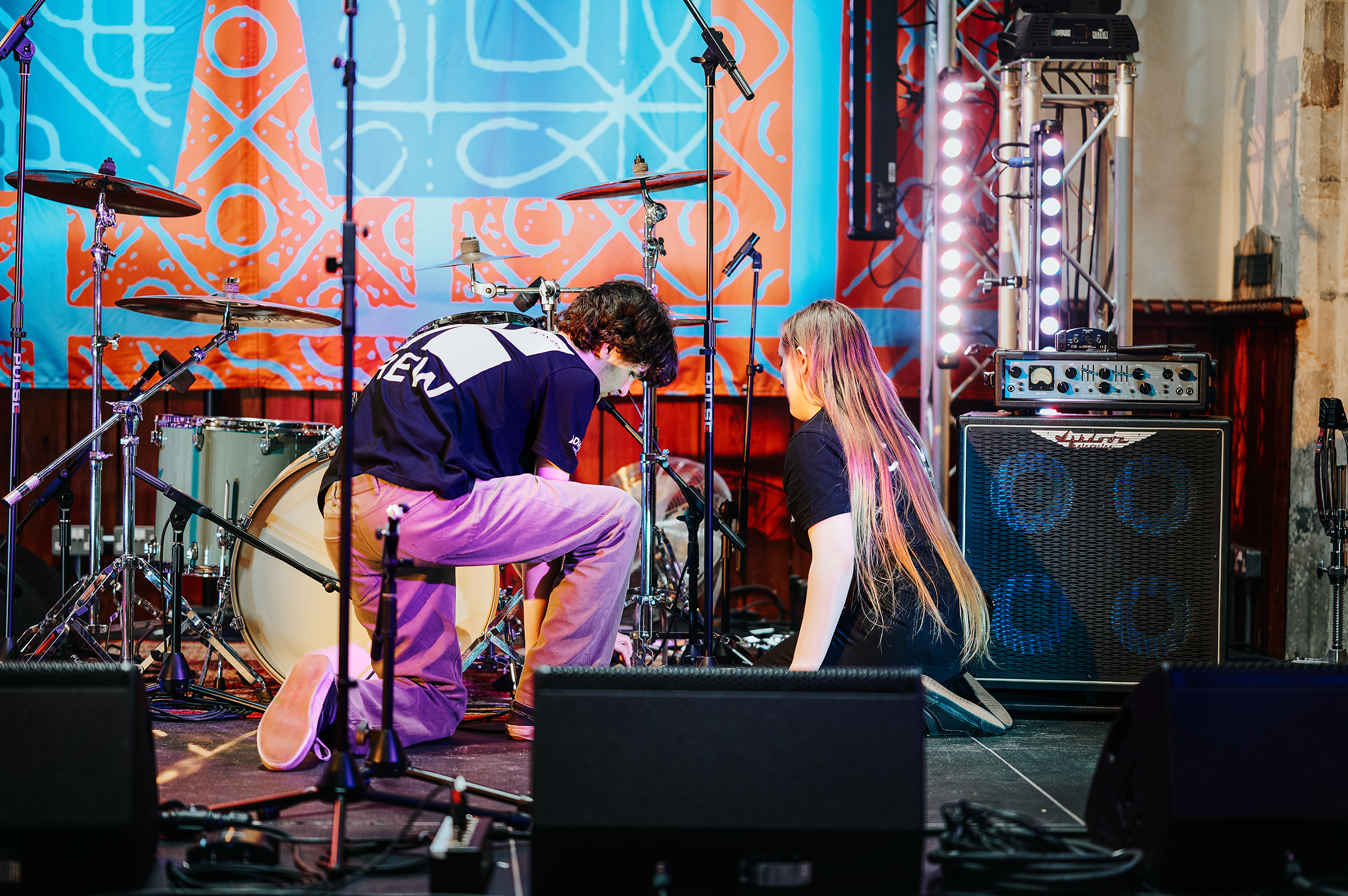 Two volunteers kneeling on stage adjusting drum equipment under colourful lighting at St Stephen’s church