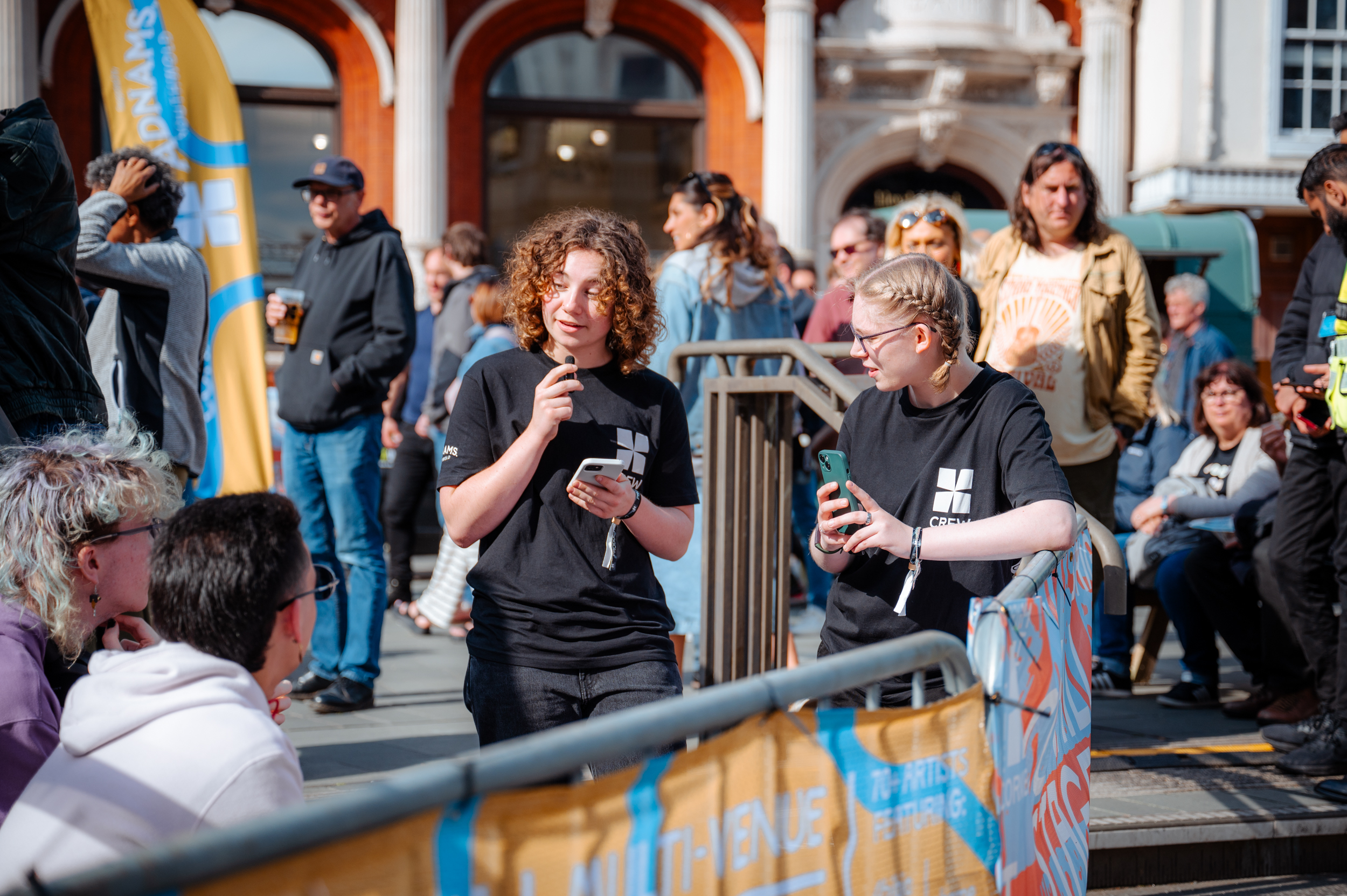 Two young volunteers wearing BTC crew t-shirts talking outside during the Cornhill stage of the festival
