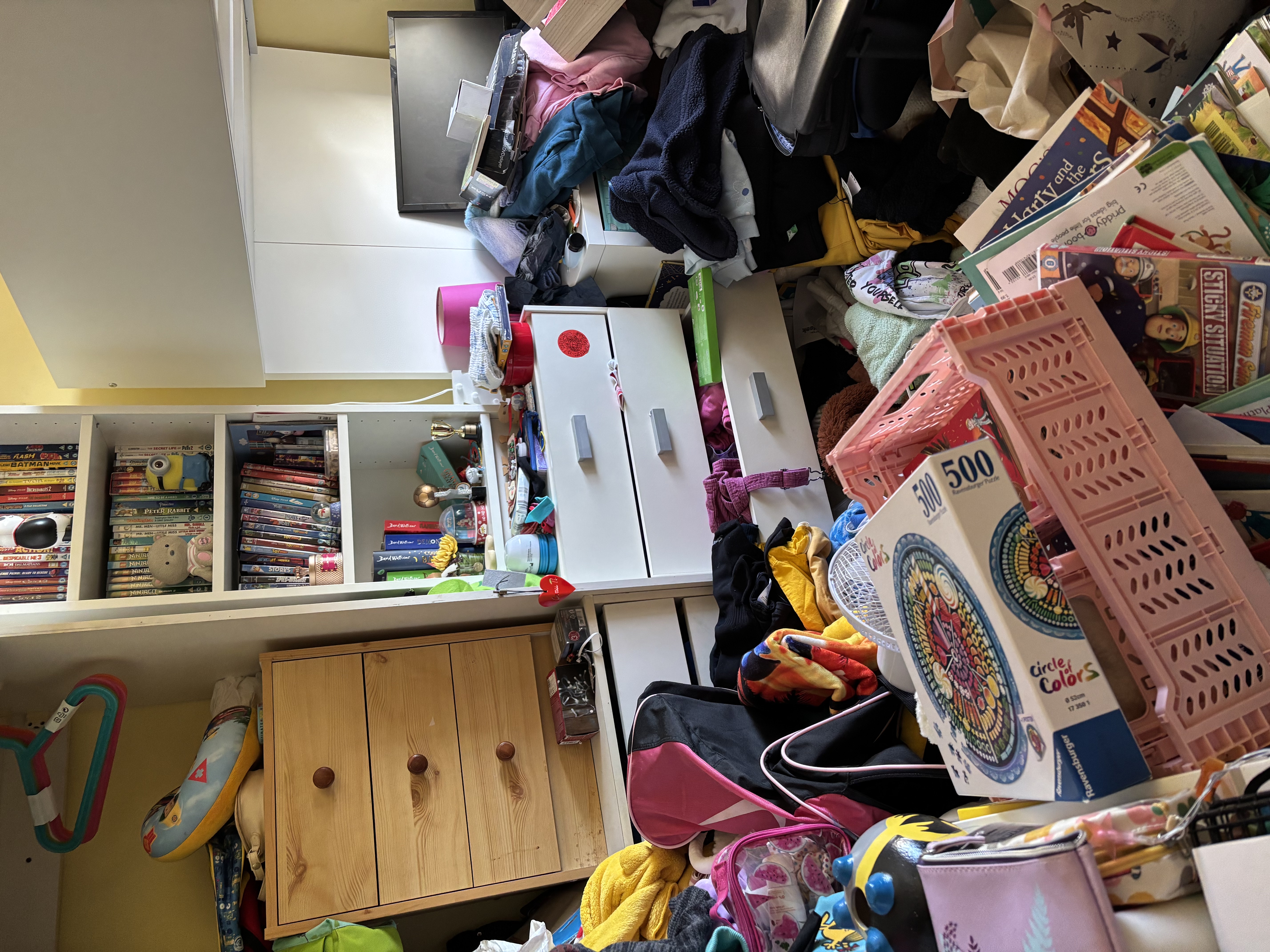 Cluttered child's bedroom with toys, clothes and belongings scattered across floor and furniture