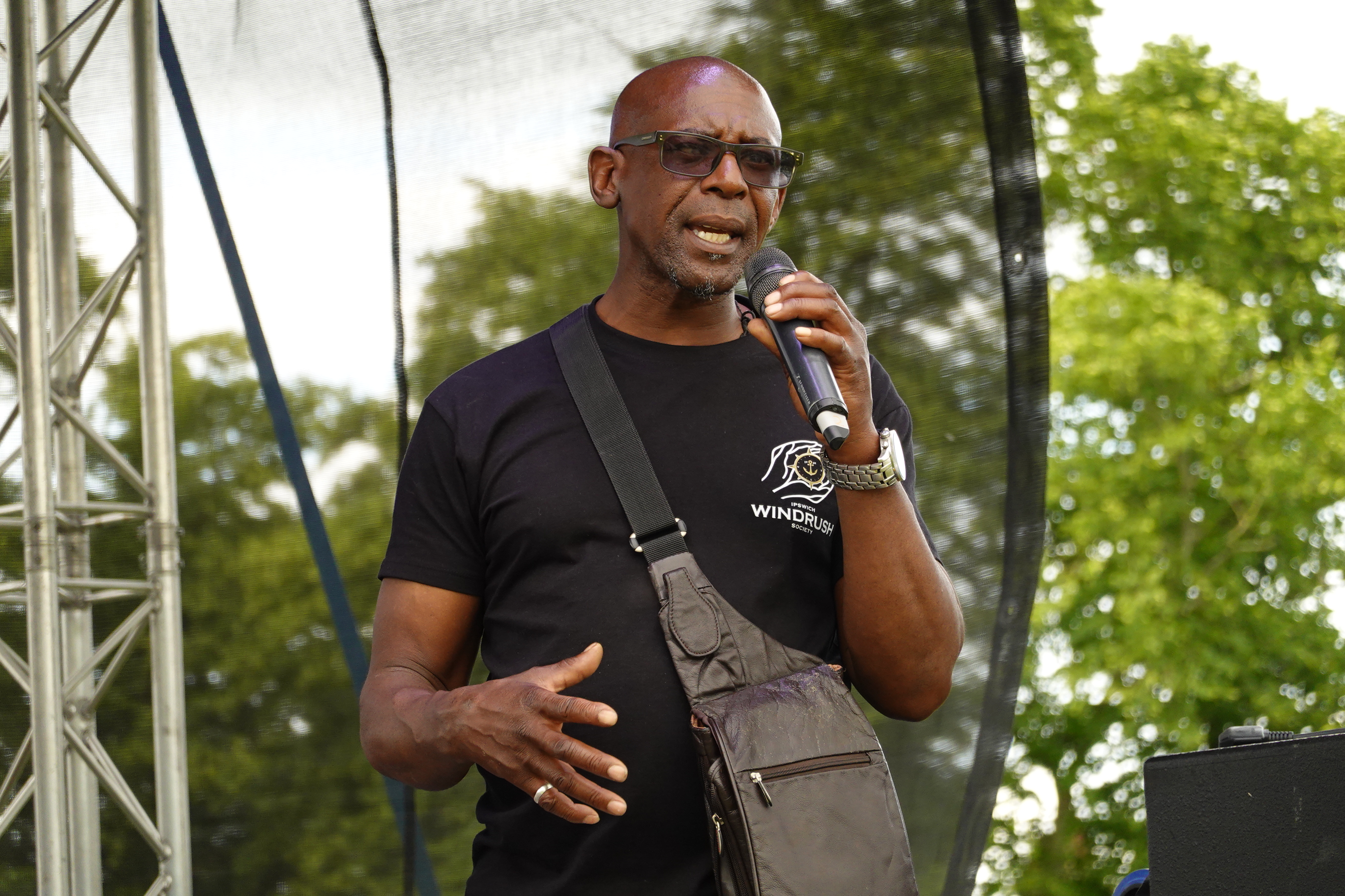Max Thomas addresses crowd from stage wearing Windrush t-shirt and holding microphone at outdoor festival