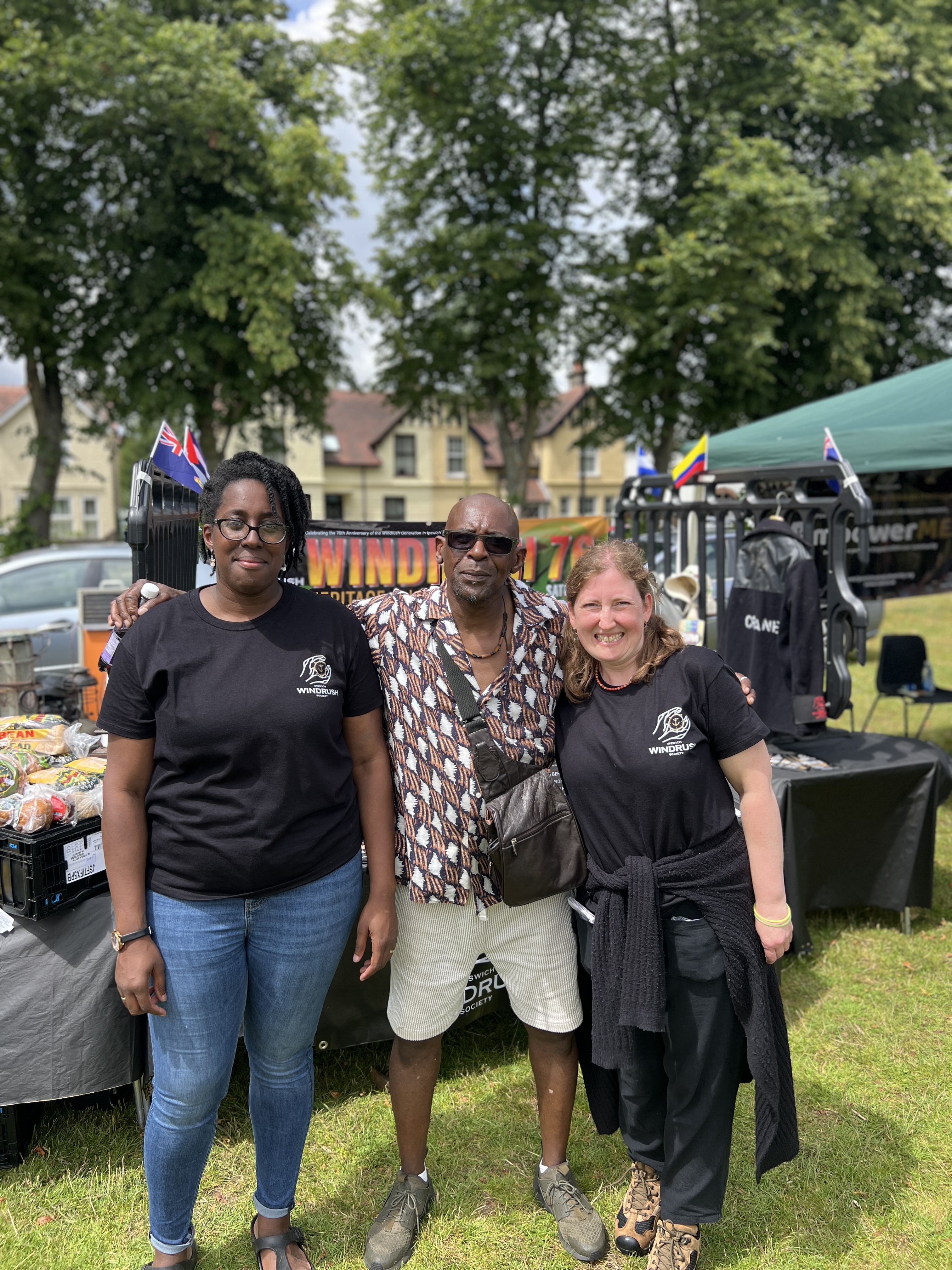 Three Windrush festival organisers pose together wearing event t-shirts with festival banners and stage visible in background