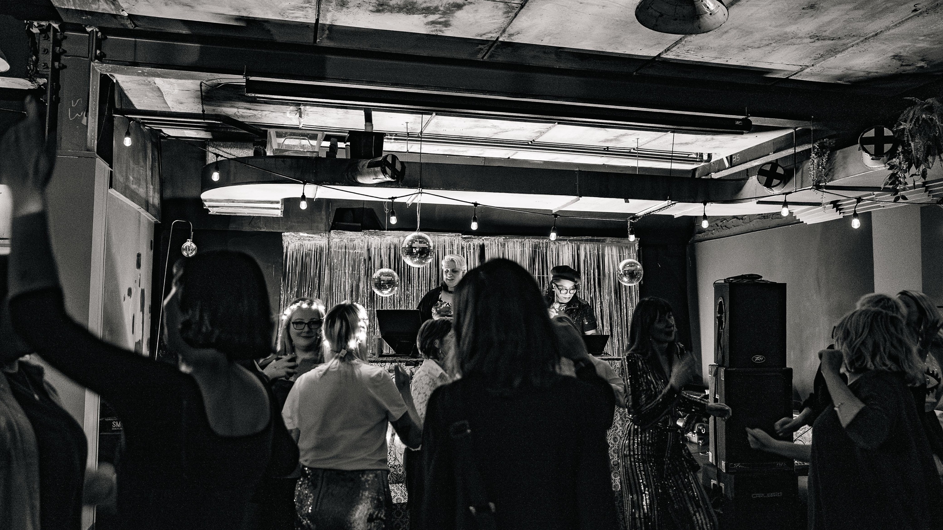 Crowd of people dancing in front of two DJs at a Grief Disco, with disco balls and silver streamers overhead