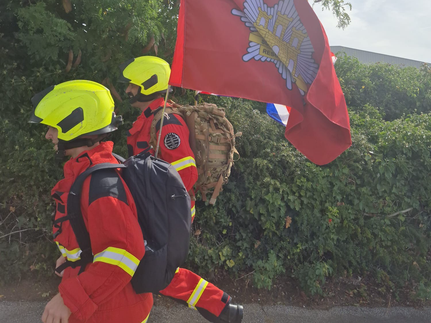 Two firefighters in protective kit jog beside a hedge carrying a large red Fire and Rescue Service flag during training for the Ipswich Half Marathon stretcher challenge