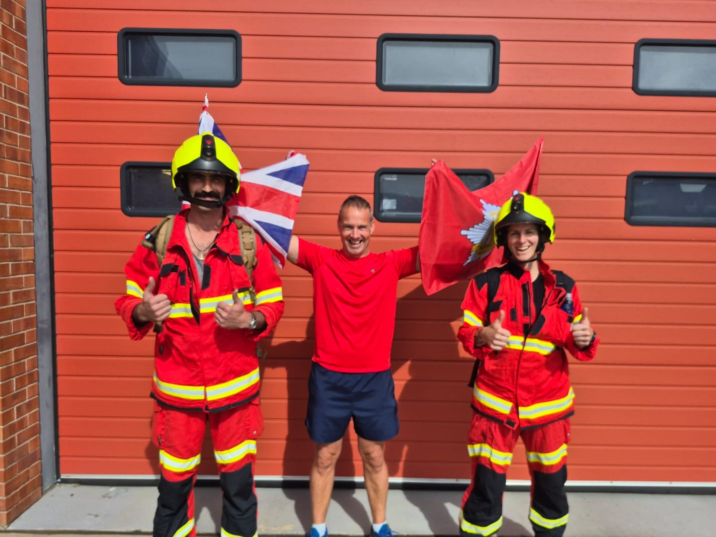 Two firefighters in full protective kit stand either side of a man in running gear holding flags, ahead of their half marathon stretcher challenge