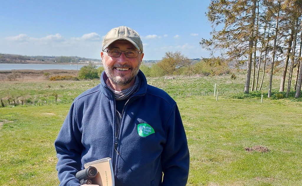 Graham Hart smiling outdoors, wearing a Suffolk Wildlife Trust jacket and cap, with trees and fields in the background