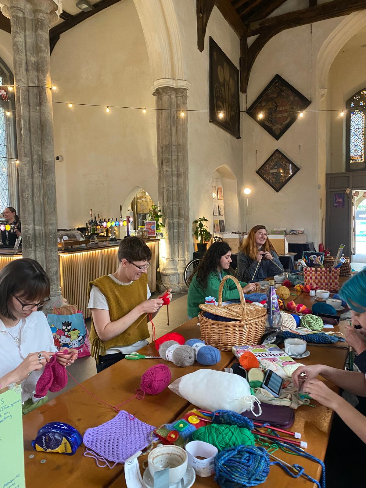 A group of people sit around a long wooden table covered with yarn and knitting supplies inside a church building, making squares for blankets