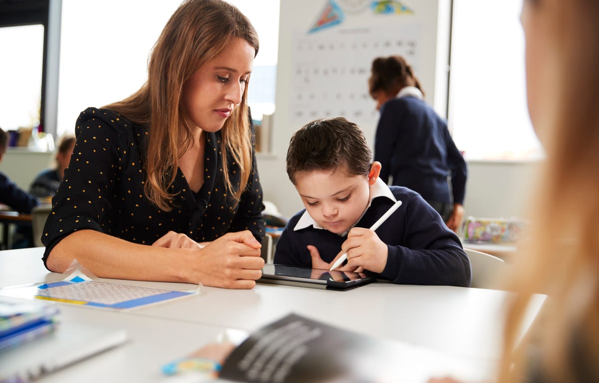 Young female teacher working with a Down syndrome schoolboy sitting at desk using a tablet computer and stylus in a primary school classroom