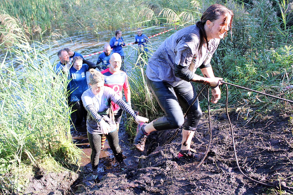 Group of Whole Hog volunteers climbing out of a muddy water pit using a rope during the obstacle race, smiling and supporting each other
