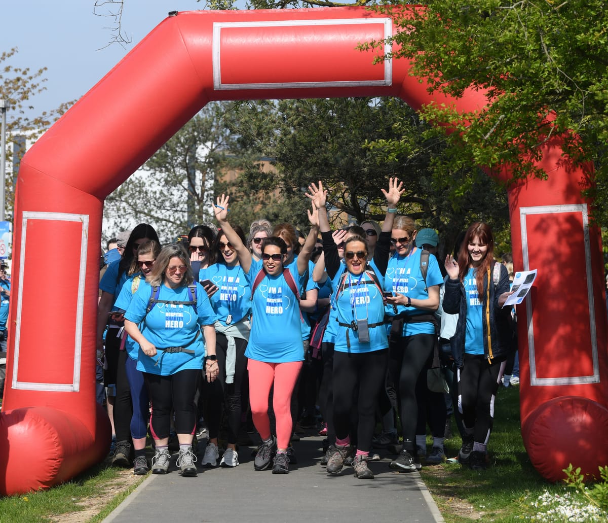 Group of walkers in blue Hospital Hero Hike t-shirts leaving the start line under a red archway