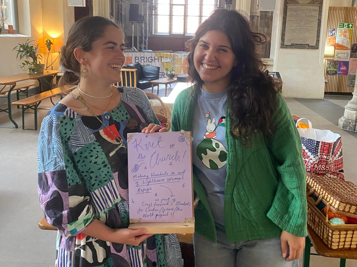 Two smiling women stand inside The Church café-bar in Ipswich, holding a hand-drawn poster for the Knit in the Church event, with knitting baskets and bags of yarn visible nearby