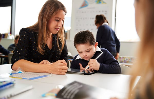 Young female teacher working with a Down syndrome schoolboy sitting at desk using a tablet computer and stylus in a primary school classroom