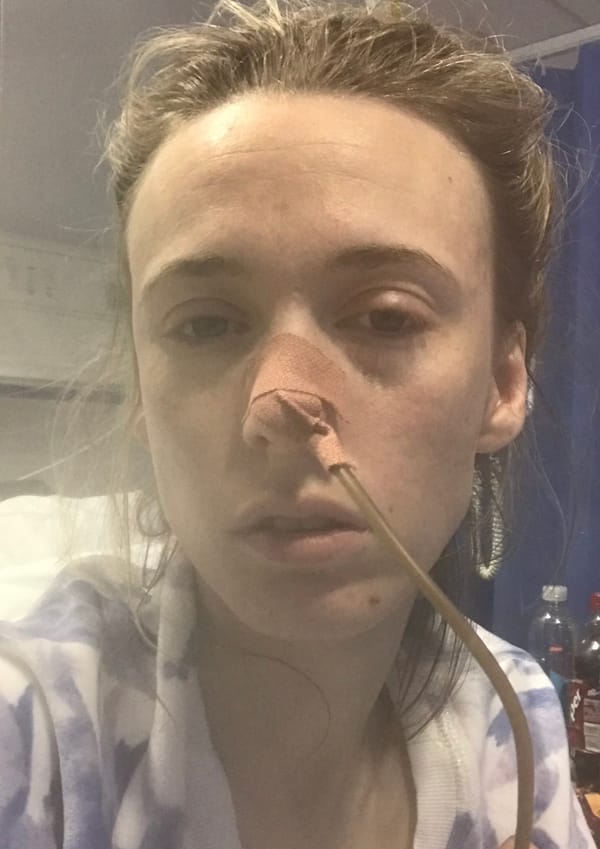 Close-up of Monica Thomas in a hospital bed with a nasogastric tube taped to her nose, looking visibly unwell and exhausted.