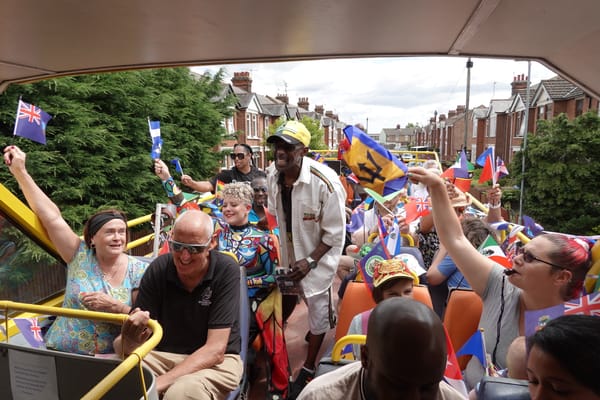 Community members of all ages wave Caribbean flags from a vintage yellow bus during Windrush festival parade through Ipswich streets