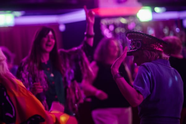 People dance under pink and purple lights at a Grief Disco, one woman wearing a sequinned disco hat
