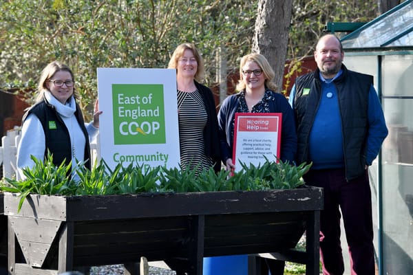 Four people standing beside a planter holding East of England Co-op and Hearing Help Essex signs