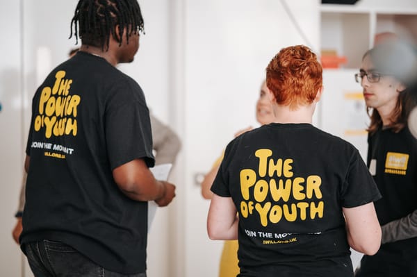 Back view of young people wearing black t-shirts with yellow text reading "THE POWER OF YOUTH" and "JOIN THE MOVEMENT #IWILL.ORG.UK"